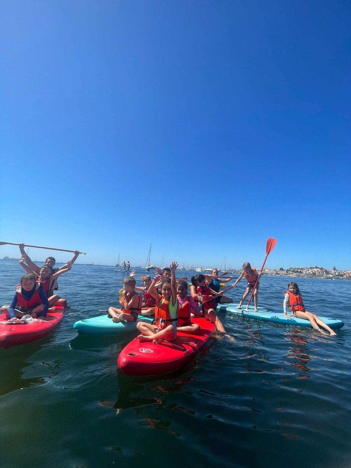 A group of students in life jackets rafted together on kayaks and paddleboards, waving on calm bay water