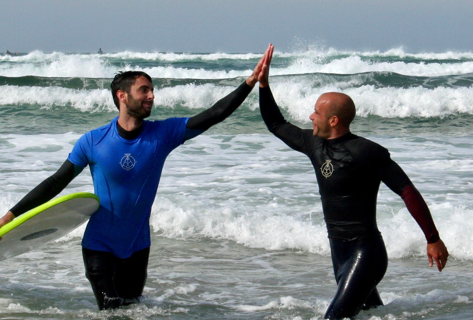 Two surfers high-fiving in the ocean wearing Cascais Routes shirts
